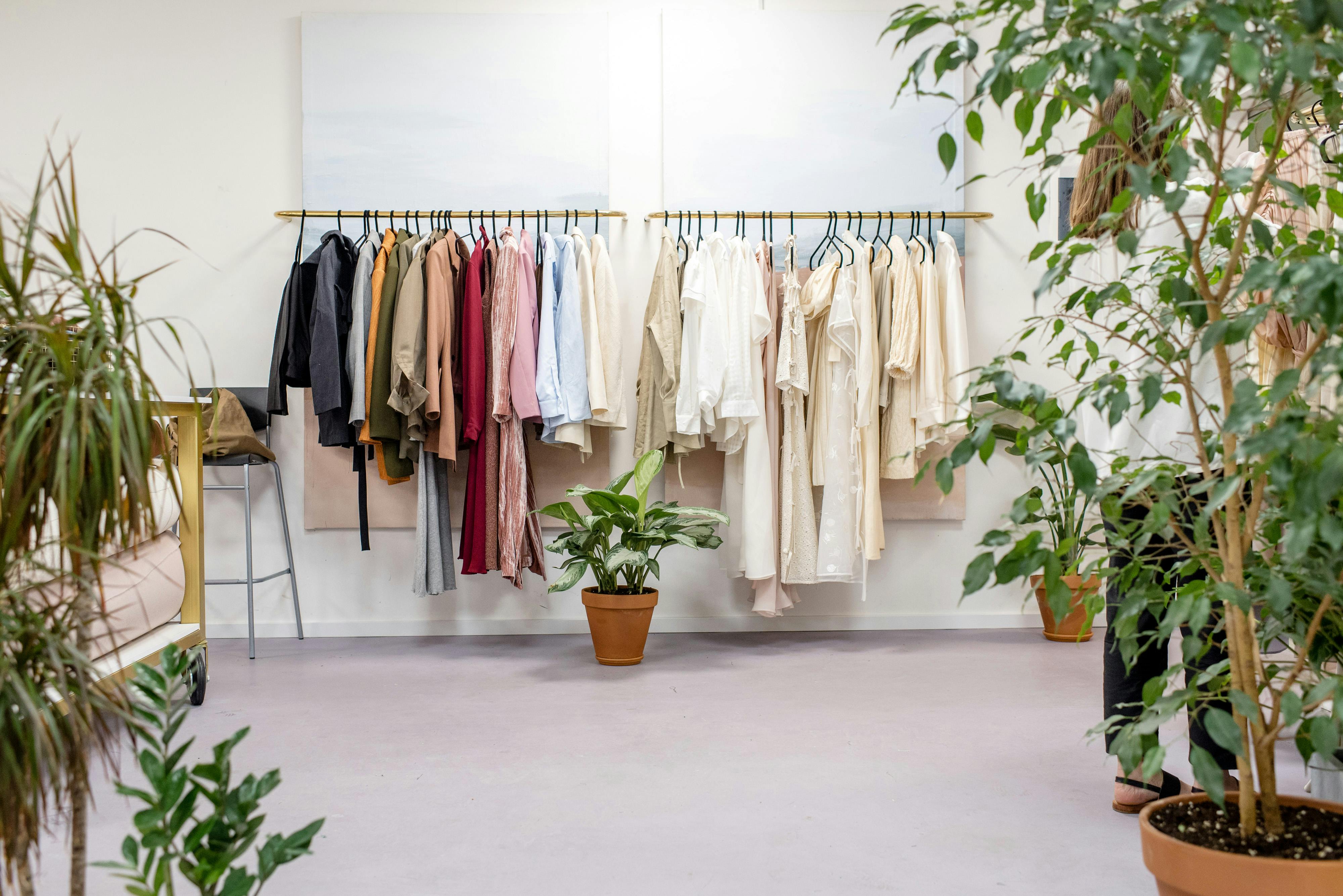 Clothing racks filled with neutral-toned garments in a minimalist boutique surrounded by indoor plants.